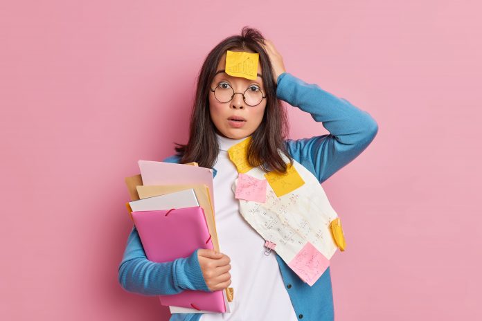 Puzzled shocked female office worker overloaded with paper work stunned to have deadline for finishing research holds folders keeps hand on head wears round spectacles poses against pink background Puzzled shocked female office worker overloaded with paper work stunned to have deadline for finishing research holds folders keeps hand on head wears round spectacles poses against pink background